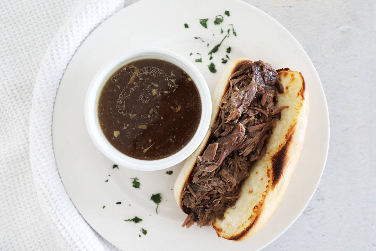 An open French dip sandwich next to a small white bowl of au jus on a white plate. 