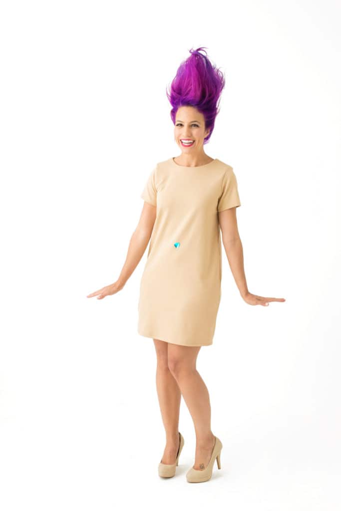 A Woman wearing a tan t-shirt dress with a jewel belly button ornament and purple hair sticking up dressed up like this for a 90's toy troll halloween costume.