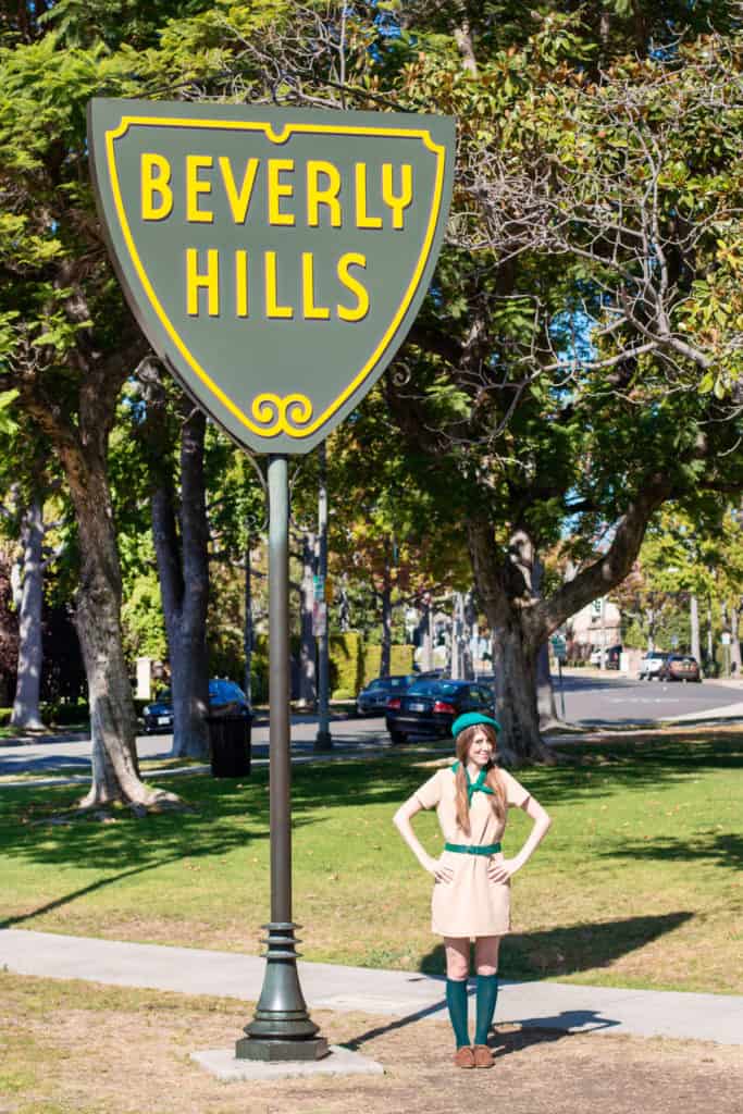 Troop Beverly Hills girl scout costume. A woman wearing a tan dress, green beret and green scarf, standing by a large Beverly Hills street sign.