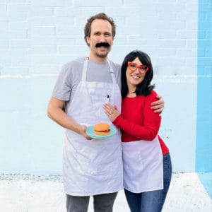 Couples Bob's Burgers costume - man wearing a gray t-shirt, busy mustache and apron with woman wearing red tshirt, red glasses, white apron and black wig.