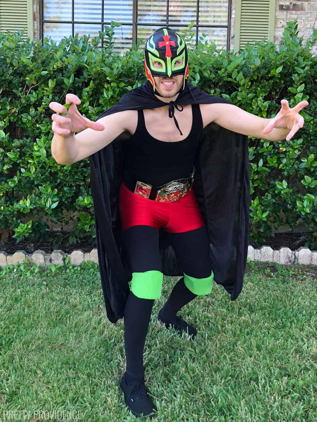 Man in a lucha libre Mexican wrestling costume, with a black, green and red luchador mask on, a black cape and black clothing. 