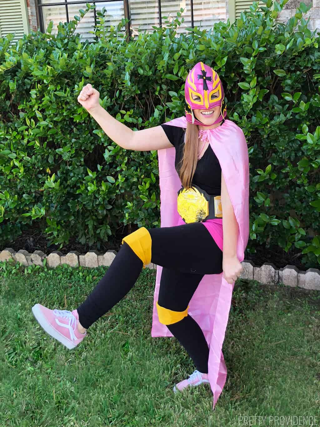Woman in a lucha libre Mexican wrestling costume with a pink luchador mask on, a pink cape, pink shorts and black clothing. 