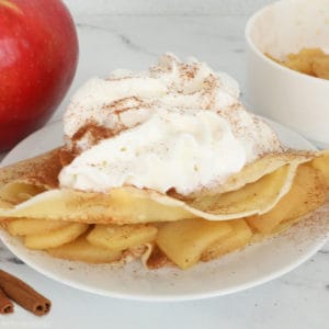 Homemade crepe filled with cinnamon apples, topped with whipped cream, and dusted with cinnamon on a white plate.