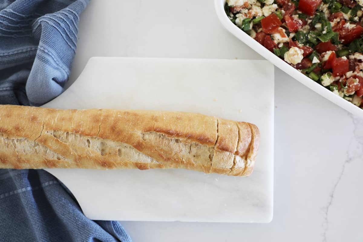 A baguette loaf on a white cutting board next to bruschetta dip. 