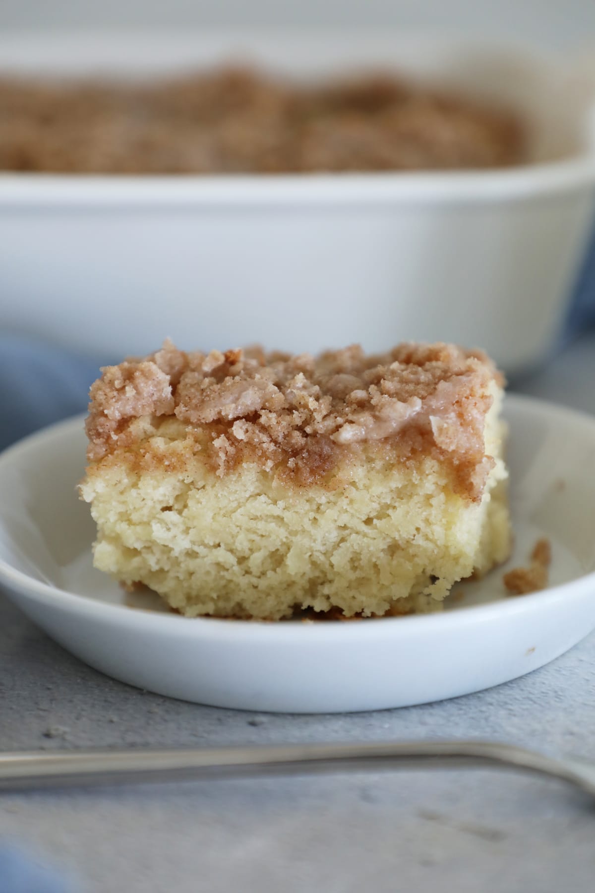 A slice of coffee cake with streusel topping and vanilla glaze in front of the pan of cake. 