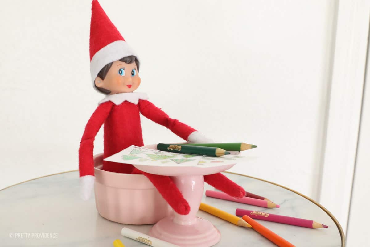 Elf on a shelf shown from the side sitting in a pink ramekin at a pink table coloring some small christmas coloring sheets with mini colored pencils. 