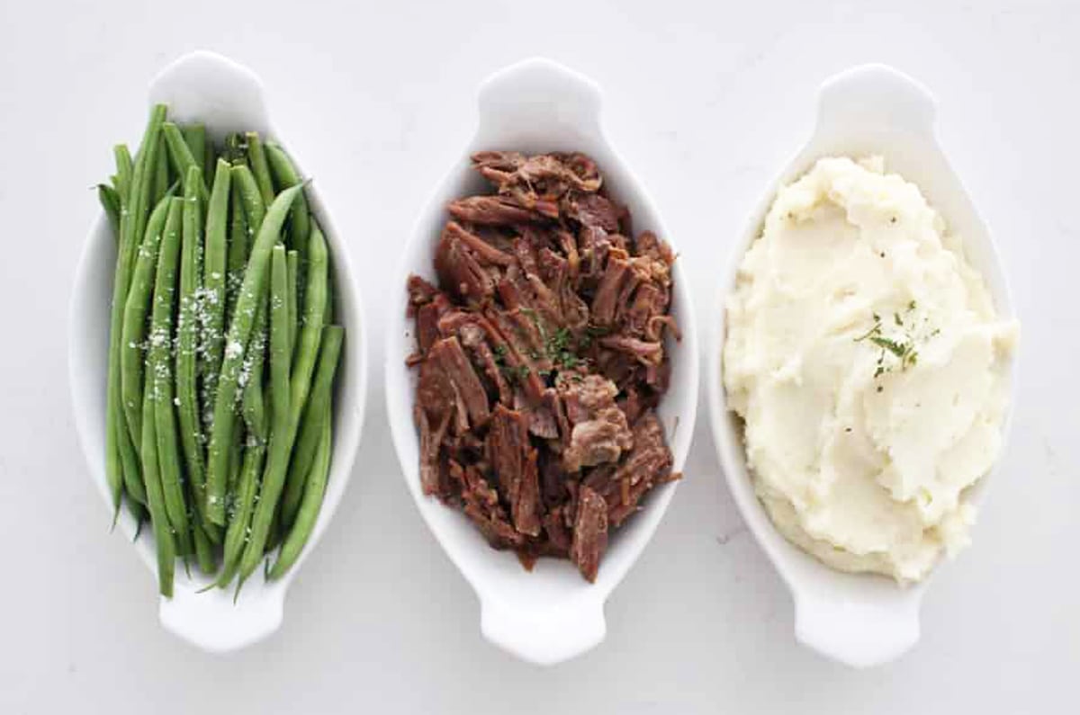 Three white dishes containing green beans, pot roast, and mashed potatoes. 
