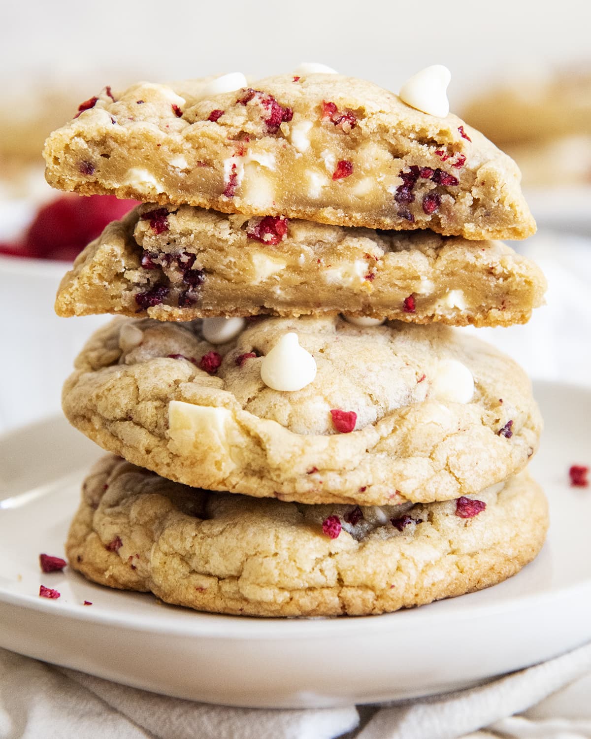 White chocolate raspberry cookies onn a plate stacked high. The top cookie is cut in half to reveal the raspberries and white chocolate chips within.