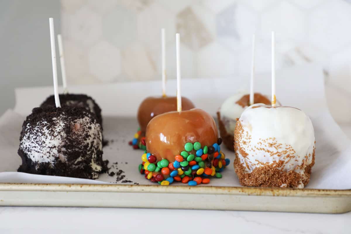 A photo of six homemade caramel apples with different toppings on a cookie sheet lined with wax paper. 