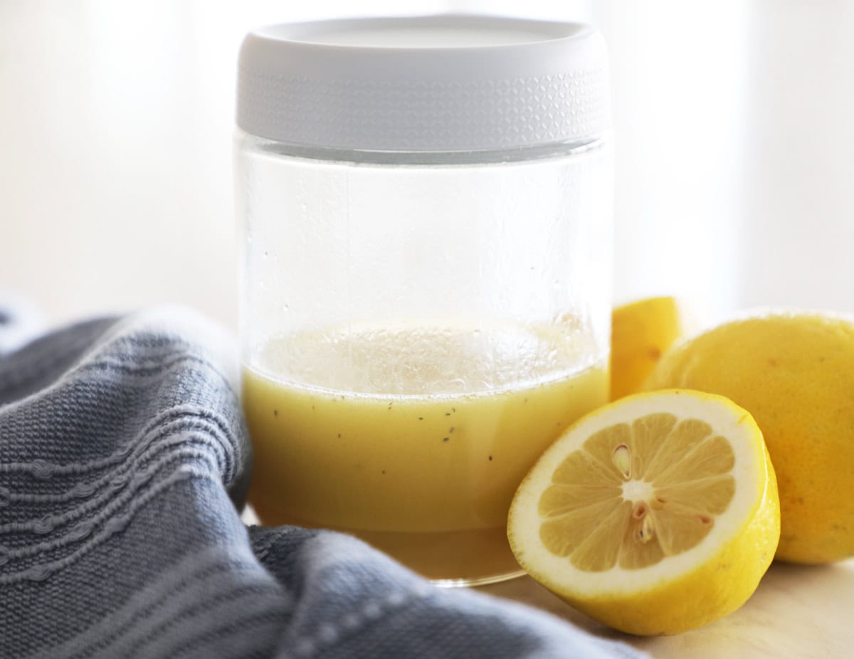 A small mason jar with a lid filled with homemade Jennifer Aniston Salad Dressing, next to lemons and a blue tea towel.