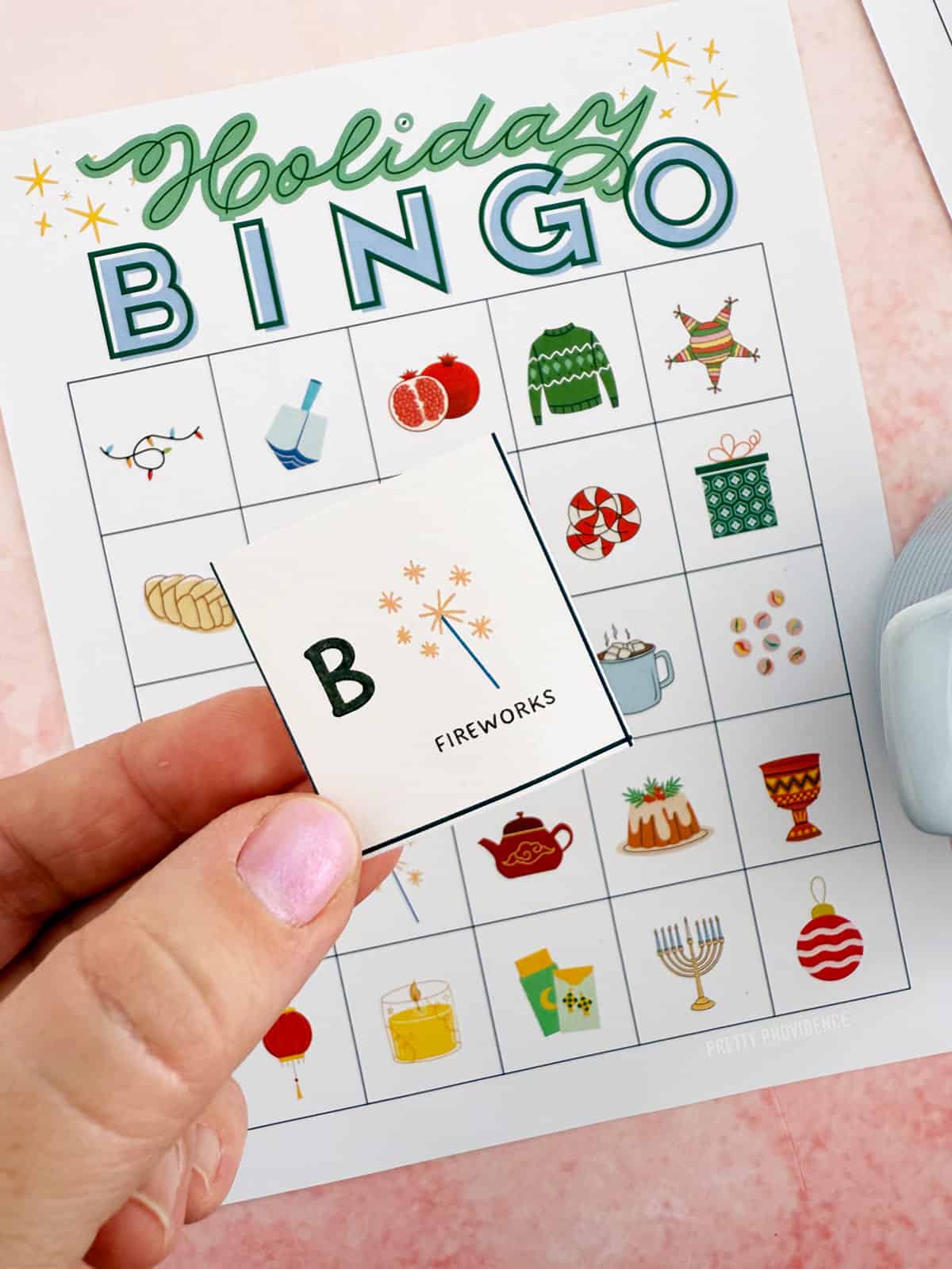 Holiday bingo card with a calling card being held over it. The calling card shows the letter 'B' and and image of fireworks on it.