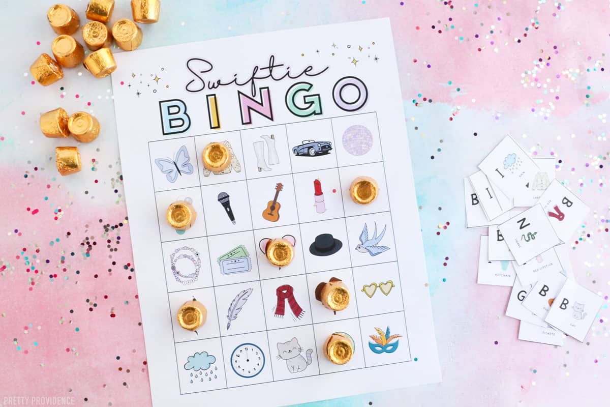 Printable Swiftie Themed Bingo Game Taylor Swift Bingo with themed pictures, on a flat lay with glitter and rolo candy on the side.